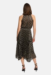 Schwarzes, plissiertes Kleid mit Halterneck, das aufwendige goldene Wellenmuster zeigt. Der Saum ist unregelmäßig und reicht bis zur Mitte der Waden, kombiniert mit Riemchen-Heels.