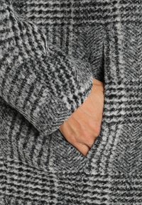 Grå och svart rutigt ullrock med en strukturerad yta, med en hand delvis synlig i en sidoficka.