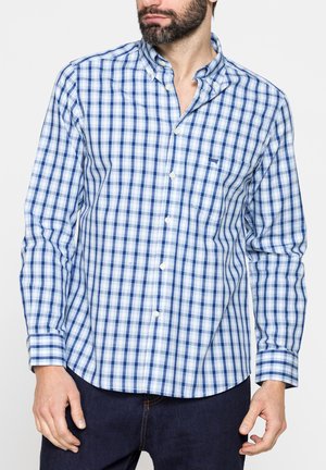 Uomo che indossa una camicia a quadri blu e bianca con maniche lunghe e jeans scuri, in piedi contro uno sfondo semplice.