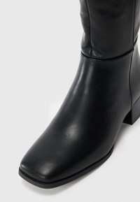 Bottine en cuir noir avec un bout carré, une texture lisse et un petit talon carré. Un détail de couture à la cheville met en valeur le design.