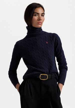 Pull col roulé en tricot marine avec un motif en chevrons, arborant un petit logo rouge. Associé à un pantalon noir taille haute et une ceinture à boucle dorée.