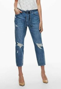 Jeans de mezclilla azul claro con un corte de talle medio, detalles desgastados y cintura con lazo. Longitud recortada y diseño de pierna recta. Combinados con tacones nude.