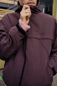 Giacca a motivi bordeaux con design a griglia, colletto alto, chiusura con zip e dettagli neri. Nota: polsino rosa chiaro visibile sotto.