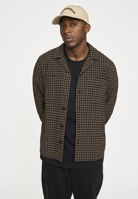 Brun og creme ternet skjortejakke med knapper, båret over en sort T-shirt. Kombineret med sorte bukser og en beige kasket.