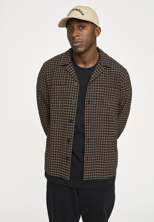 Brun og creme ternet skjortejakke med knapper, båret over en sort T-shirt. Kombineret med sorte bukser og en beige kasket.