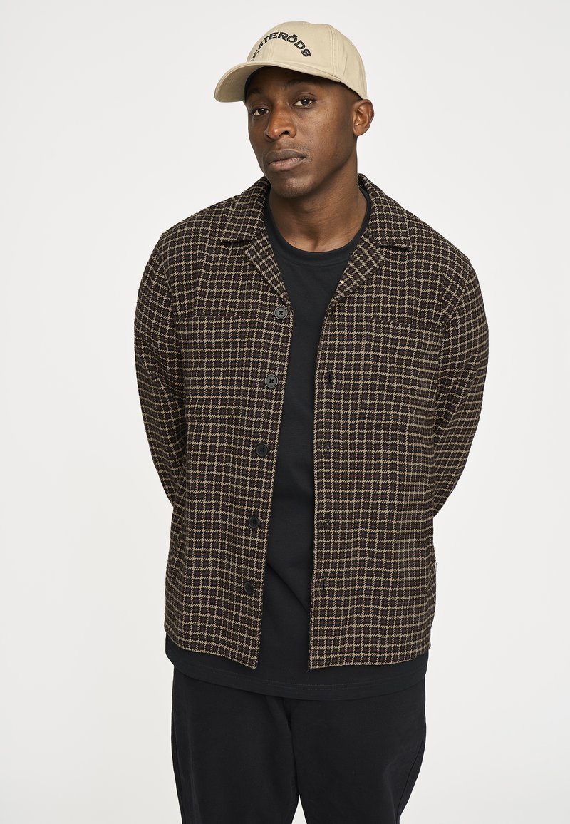 Brun og creme ternet skjortejakke med knapper, båret over en sort T-shirt. Kombineret med sorte bukser og en beige kasket.