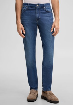 Mężczyzna ubrany w niebieskie jeansy o prostym kroju i beżowe zamszowe mokasyny, stojący na jednolitym jasnym tle.