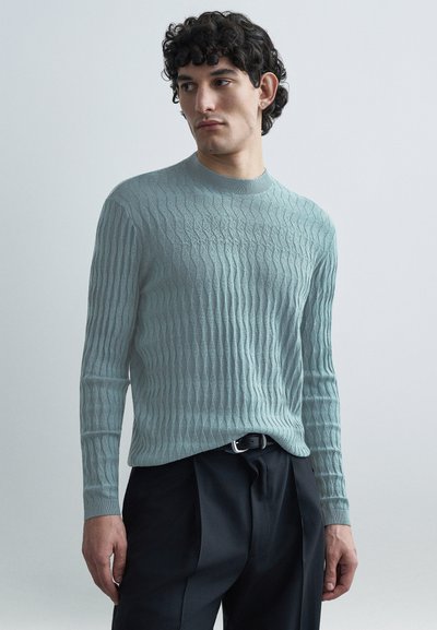 Pull tricoté bleu clair à motifs avec un col rond et des manches longues, présentant un motif ondulé texturé, associé à un pantalon foncé.