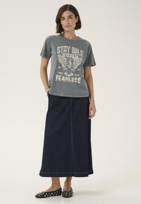 Graues Grafik-T-Shirt mit Adlerdruck, kombiniert mit einem langen, dunklen Jeansrock. Schwarze Ballerinas, verziert mit silbernen Nieten, vervollständigen das Outfit.