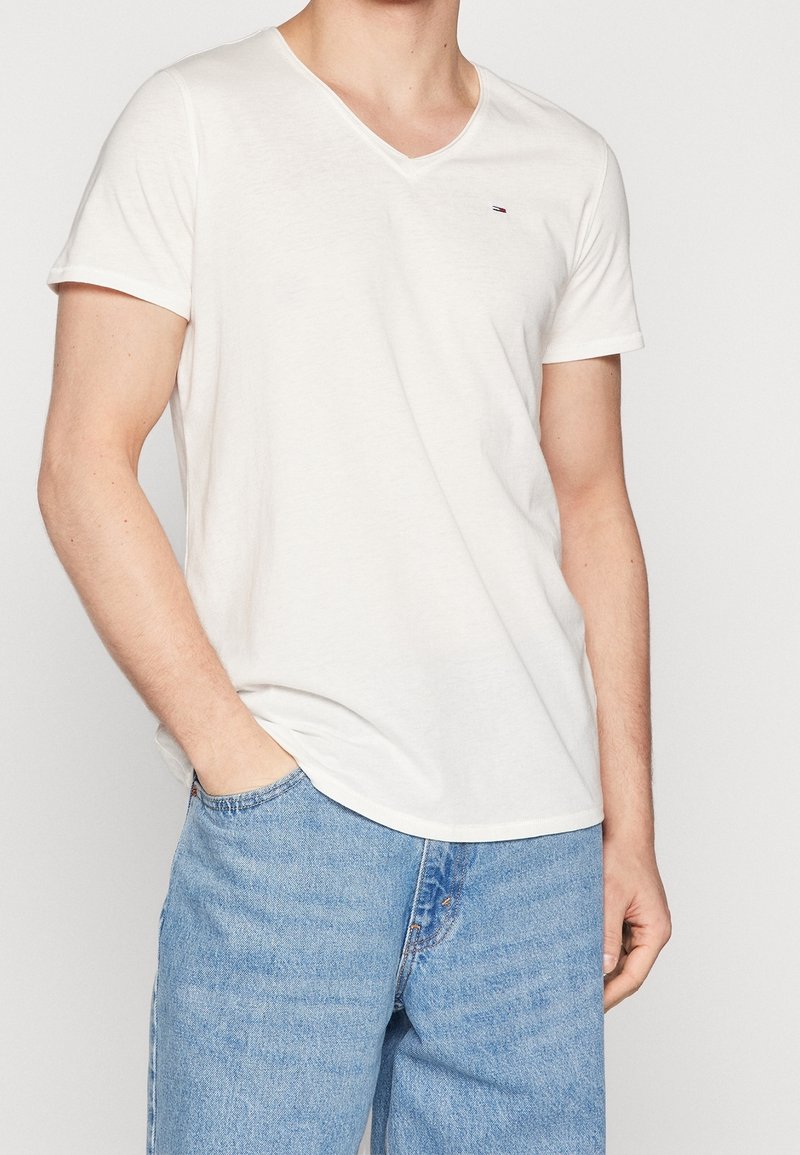 T-shirt blanc à manches courtes en col en V, fabriqué en coton doux, présentant un petit logo sur la poitrine et une coupe décontractée. Porté avec un jean bleu.
