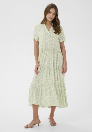 Kobieta ubrana w jasną zieloną sukienkę midi w wzory z krótkimi rękawami i beżowe sandały na obcasie, stojąca na tle białego, jednolitego tła.