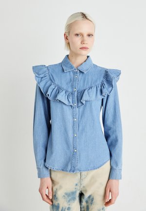 Chemise en denim bleu clair avec un col et des épaules à volants, devant boutonné, manches longues et ourlet arrondi. Portée avec un pantalon tie-dye.