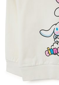 Sudadera blanca con un diseño gráfico de un conejo saltando sobre un texto colorido "cinnamoroll", con una textura de tela suave y puños acanalados.