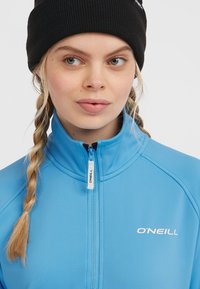 Gaiši zila ar rāvējslēdzēju kabata ar augstu apkakli, ar baltu "O'Neill" logotipu un strukturētu audumu. Apvienota ar melnu ribota tipa cepuri.