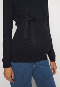 Czarny sweter z dzianiny o teksturowanym wzorze, związywany w talii i długimi rękawami. W połączeniu z niebieskimi dżinsami.