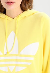 Gul hoodie tillverkad av mjukt material, med en stor vit logotypdesign med tre ränder och ett bladmönster. Dragsnören i matchande gul.