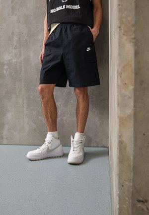 Schwarze Cargo-Shorts mit einer Vorderseite-Tasche und elastischem Bund, kombiniert mit weißen Nike High-Top-Sneakern und weißen Socken. Glatte Textur.