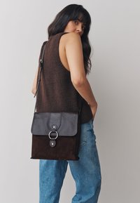 Geantă crossbody din piele și piele întoarsă, de culoare maro închis, cu un finisaj texturat, accesorii argintii și o închidere cu clapetă care are un detaliu sub forma unui inel circular.