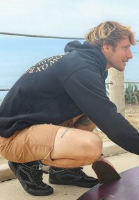 Haut à capuche noir avec texte doré, shorts beige et baskets noires. L'individu est accroupi près d'une planche de surf sur une surface en béton.