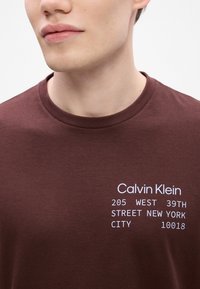Camiseta de manga corta color burdeos con texto impreso en blanco: "Calvin Klein 205 WEST 39TH STREET NEW YORK CITY 10018," cuello redondo, textura suave.