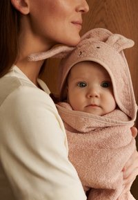 Różowy ręcznik dla niemowląt z frotté z uszami królika, owinięty wokół dziecka. Skupienie na miękkiej fakturze i zaokrąglonych kształtach. Jasnobrązowe tło.