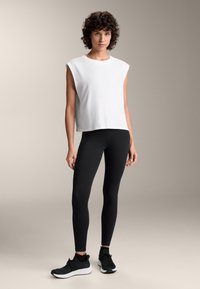 Weißes ärmelloses Top aus leichtem Stoff, kombiniert mit schwarzen Leggings und schwarzen Sportschuhen. Einfaches Design und einfarbige Farben.