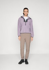 Fleece pulóver vo svetloj fialovej farbe s čiernym zipsom na golieri a vreckom na hrudi; kombinovaný s béžovými nohavicami a čiernymi topánkami. Hladká textura.