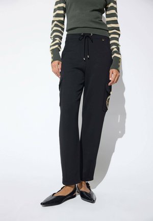 Persona con pantalones cargo negros con bolsillos laterales y zapatos planos negros puntiagudos, combinados con una camiseta de manga larga a rayas verdes y beige.