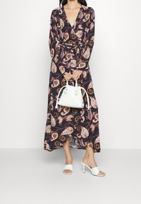 Žena v dlouhých černých šatech s paisley vzorem, držící bílou kabelku a oblečená v bílých mules na podpatku, na jednobarevném pozadí.