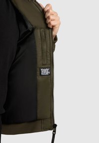 Jachetă de culoare verde măslinie cu fermoar pe față, căptușeală neagră, manșete elastice și un label cu logo în interior. Materialul pare neted și rezistent la apă.