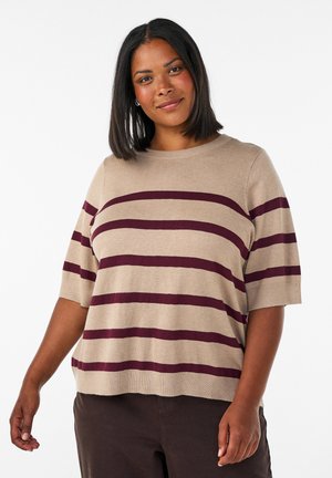 Vrouw met zwart haar tot op de schouders, gekleed in een beige sweater met korte mouwen en donkerrode horizontale strepen, staand tegen een witte achtergrond.