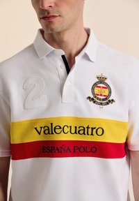 Camisa polo blanca con cuello navy, que presenta rayas horizontales rojas y amarillas. Logo bordado y el número "2" en el pecho.