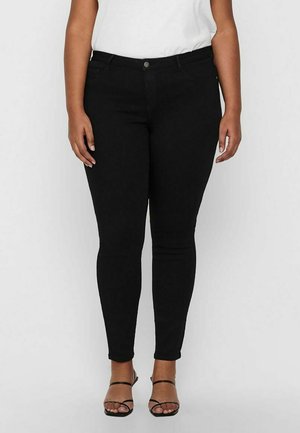 Woman wearing black fitted jeans and black strappy open-toe sandals, standing against a plain light background.