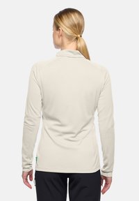 Vrouw met blond haar in een paardenstaart, gekleed in een lichtbeige lange mouw top en zwarte broek, gezien vanaf de achterkant tegen een witte achtergrond.