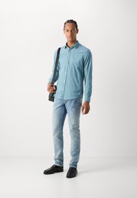 Camisa de botones de manga larga azul claro con un pequeño logo, combinada con jeans azules desgastados y zapatos negros sin cordones, llevada con una bolsa de mensajero negra.