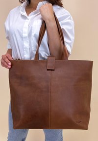 Sac fourre-tout en cuir marron avec deux larges anses, une fermeture centrale par sangle, et une texture lisse. Présente des coutures discrètes et un logo.