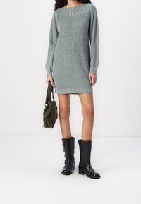 Robe pull en tricot gris avec des manches longues, un large décolleté et un ourlet côtelé. Associée à des bottes noires jusqu'au genou et un sac vert.