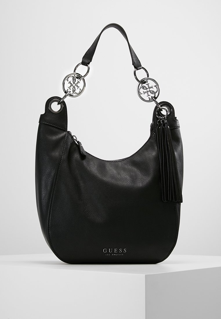Sac à épaule en cuir noir Guess avec accents logo argentés, détail pompon et poignée unique sur une surface blanche.