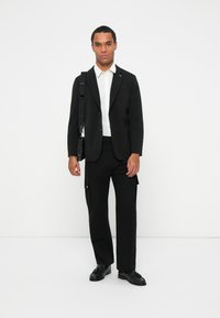 Sort skræddersyet blazer over en hvid skjorte med knapper, parret med sorte cargobukser. Sorte sko og en skuldertaske fuldender outfittet.