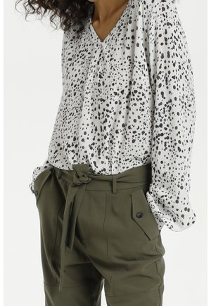 Blouse blanche à manches longues avec des taches noires irrégulières, rentrée dans un pantalon taille haute vert olive avec ceinture nouée et poches boutonnées.