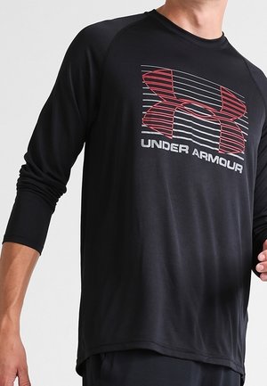 Mężczyzna w czarnej, długorękawowej sportowej koszulce z czerwonym i białym logo Under Armour oraz napisem na piersi.