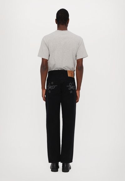 Pantalones de mezclilla negros con corte recto, que presentan costuras blancas en contraste, acentos de estrellas y un parche de cuero marrón en la cinturilla. Camiseta gris de cuello redondo.