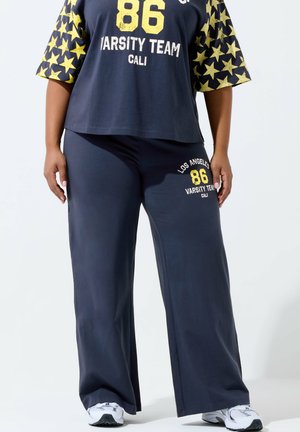 Kobieta ubrana w granatowy strój varsity z rękawami w żółte gwiazdki i białe trampki, stojąca na białym tle.