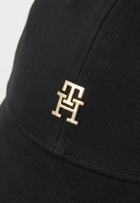 Juodas medvilninis kepurė su tekstūruota paviršiumi, priekyje turi auksinį metalinį logotipo akcentą.