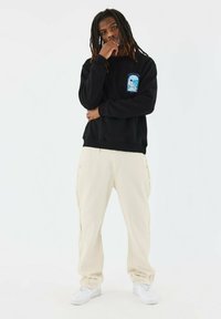 Sudadera negra con parche gráfico azul, pantalones cargo crema con bolsillos laterales y zapatillas blancas. Tejido suave y corte relajado.