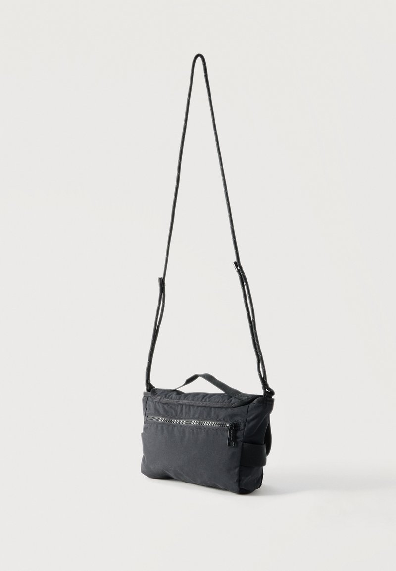 Zwarte crossbodytas met verstelbare lange schouderriem, voorvak met rits en klein handvat bovenaan, getoond tegen een effen lichte achtergrond.