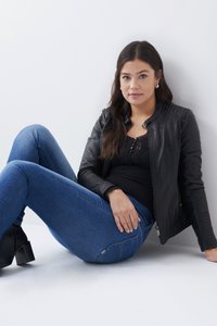 Chaqueta de cuero negra con acentos acanalados, top de encaje negro y jeans ajustados azules. La modelo se sienta sobre un fondo blanco con botas negras hasta el tobillo.