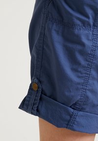 Pantaloncini in cotone blu navy con orlo arrotolato, caratterizzati da un bottone marrone. Tessuto liscio con dettagli cuciti ai lati.