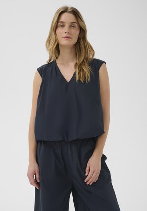 Femme aux longs cheveux châtain clair portant un haut sans manches bleu marine foncé à col en V et un pantalon ample assorti, debout devant un fond uni.