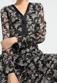 Czarna sukienka w kwiaty z przezroczystymi długimi rękawami, dekoltem w serek, skomplikowanym jasnym wzorem kwiatowym oraz jednolitymi czarnymi akcentami na dekolcie i mankietach.